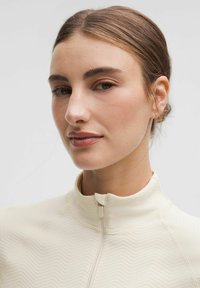 Jachetă crem cu fermoar, cu un model texturat în zigzag. Dispune de un guler înalt și un design minimalist.