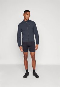 Muškarac u tamnoj biciklističkoj jakni na zatvaranje, crnim biciklističkim kratkim hlačama, crnim čarapama i crnim biciklističkim cipelama na jednobojnoj pozadini.