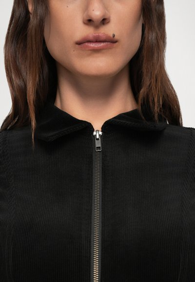 Primer plano del rostro inferior y el cuello de una mujer que lleva una prenda negra con cremallera, con una cremallera plateada visible y una marca de belleza encima del labio superior.