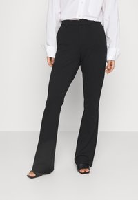 Kobieta ubrana w czarne, wysokie na talii, rozszerzane spodnie, białą, długą koszulę i czarne, otwarte na palce, kwadratowe obcasy na jednolitym tle.