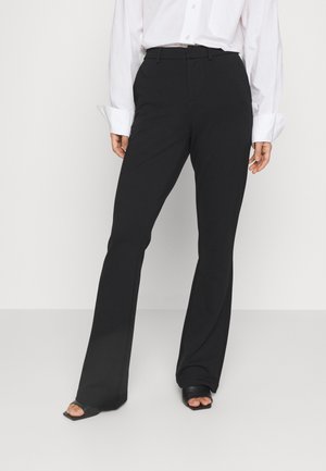 Donna che indossa pantaloni neri a vita alta e svasati, una camicia bianca a maniche lunghe e scarpe nere aperte con tacco quadrato su uno sfondo neutro.