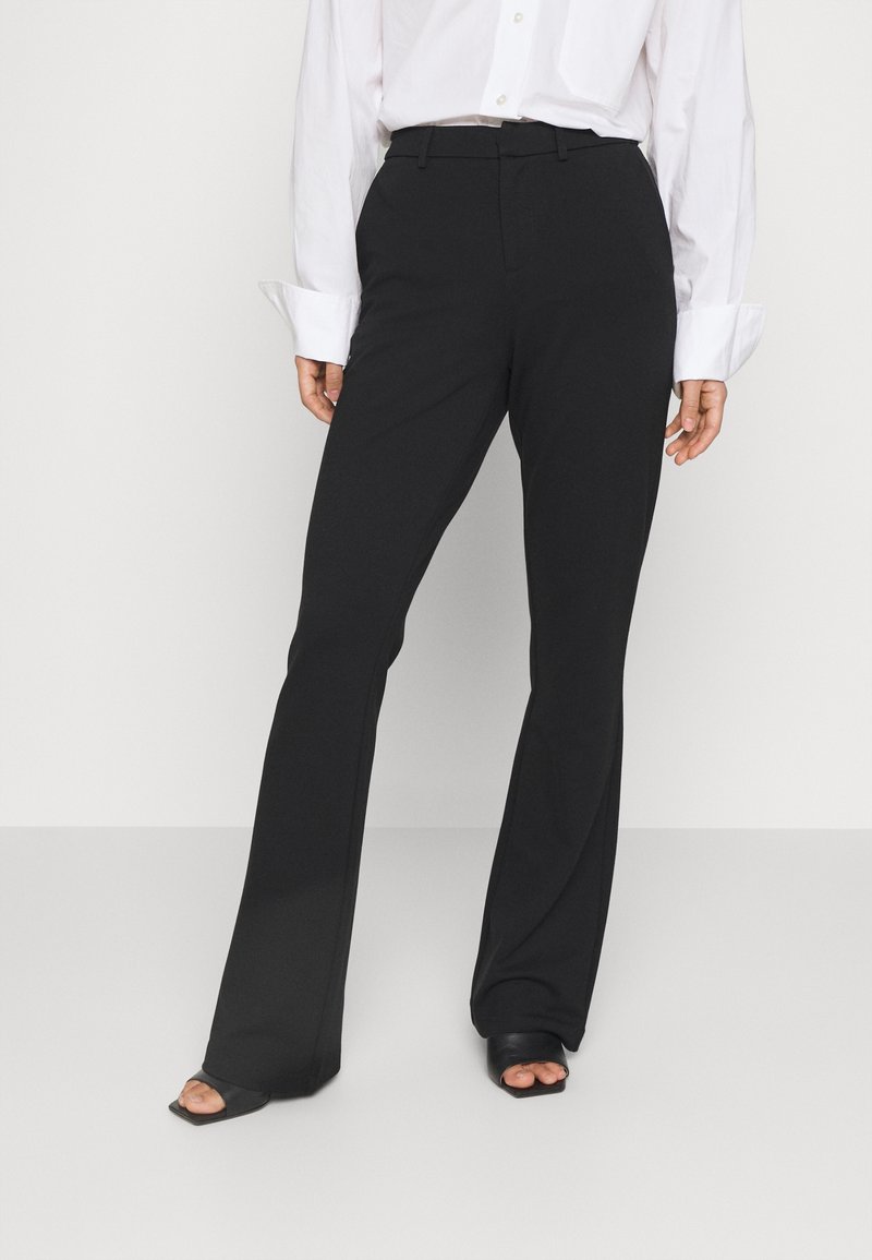 Kobieta ubrana w czarne, wysokie na talii, rozszerzane spodnie, białą, długą koszulę i czarne, otwarte na palce, kwadratowe obcasy na jednolitym tle.