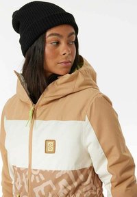 Giacca zip-up beige e bianca con cappuccio, caratterizzata da un motivo geometrico nella parte inferiore e un piccolo logo. Berretto di maglia nera indossato sulla testa.