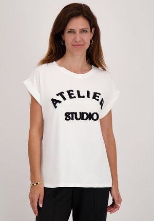 Een vrouw draagt een wit T-shirt met korte mouwen met "ATELIER STUDIO" in zwarte letters, zwarte broek, gouden ringoorbellen en een armband.