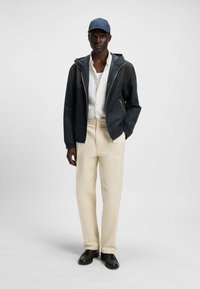 Chaqueta azul marino, camisa blanca de botones, pantalones crema y zapatos negros, acompañados de una gorra azul. Texturas suaves y un diseño ajustado.