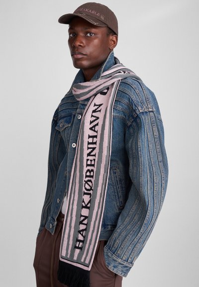 Vilnas šalle ar vertikālām svītrām rozā un pelēkā krāsā, ar melniem burtiem "HAN KJØBENHAVN." Malas apdare ar frizēm un ģērbta virs džinsu jakas.