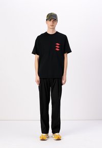Svart T-skjorte med kort arm og rødt grafisk design på brystet, sammen med svarte bukser og sneakers med gule detaljer. Kamuflasjert caps på hodet.