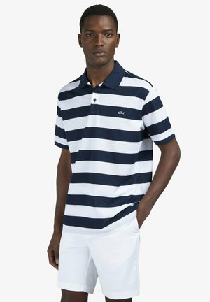 Homme portant un polo à rayures horizontales bleu marine et blanches et un short blanc, debout avec une main dans la poche sur fond uni.