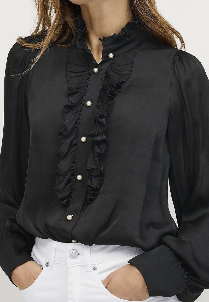 Blouse en soie noire avec un devant à volants, des boutons en perle, un col montant et des manches longues avec poignets froncés, portée avec un pantalon blanc.