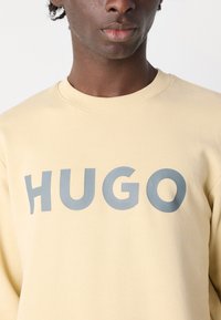Lysegul hettegenser med stor grå "HUGO" tekst på forsiden. Laget av mykt stoff med rund hals og ribbestrikkede mansjetter.