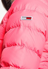 Růžová nafouklá bunda s prošívanými částmi a našitou nášivkou v námořnické modré, červené a bílé barvě na rameni. Hladká, lesklá textura látky.