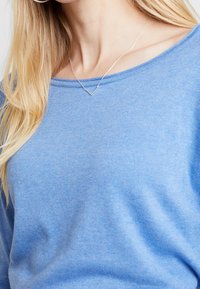 Ljusblå tröja med mjuk textur, rund halsringning och ribbad mudd. Har ett svalt silverhalsband i V-form.