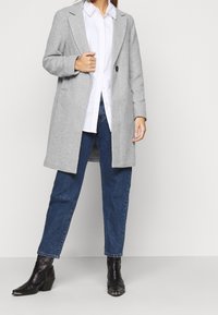 Nainen, jolla on harmaa villakangastakki, valkoinen paita, siniset farkut ja mustat nilkkurit, seisoo vaaleaa taustaa vasten.