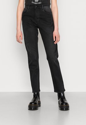 Persoon draagt zwarte high-waisted rechte jeans en zwarte plateau enkellaarsjes, staand tegen een effen lichte achtergrond.