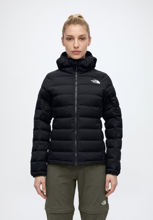 Une femme porte une doudoune noire à capuche North Face et un pantalon olive sur un fond blanc uni.
