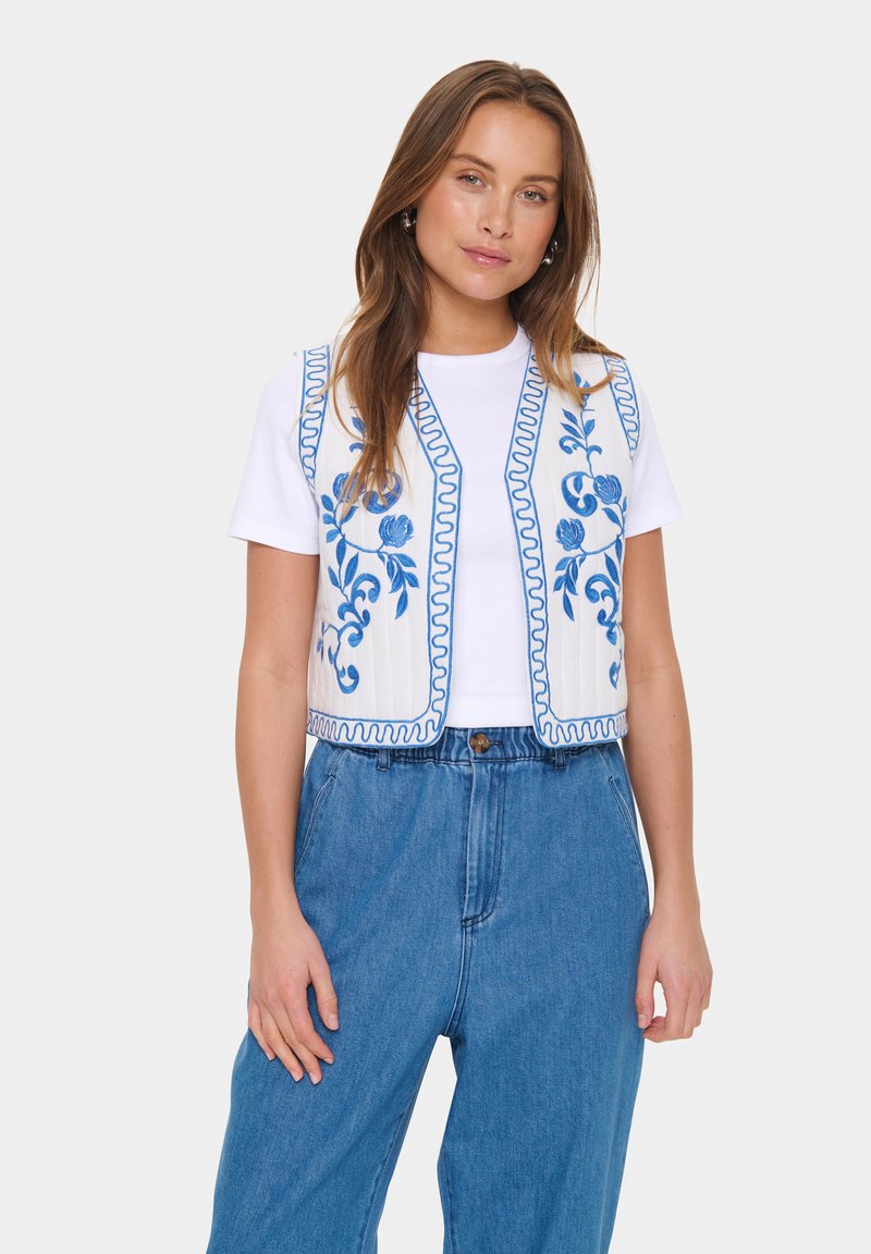 Vitt ärmlöst linne med blå blommönster och scallopade kanter, i kombination med en vit t-shirt och högmidjade blå jeans.
