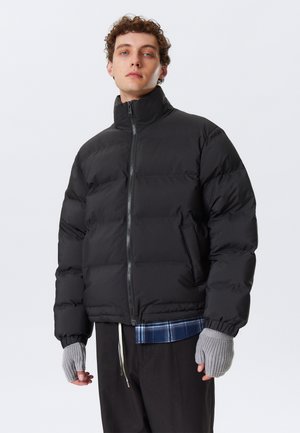 Sort puffer-jakke med høj krave, lynlås og sidelommer. Modellen har grå fingerløse handsker på over en blå ternet skjorte.