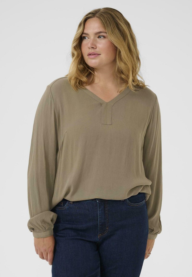 Khakifarbene Bluse mit langen Ärmeln, V-Ausschnitt und dezenter Textur. Mit gerafften Manschetten und lässiger Passform, kombiniert mit dunklen Jeans.
