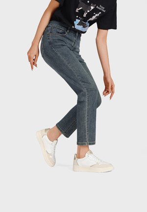 Persona che indossa jeans scuri corti, sneaker bianche e una t-shirt nera con illustrazioni di cani, piegata leggermente in avanti su uno sfondo semplice.