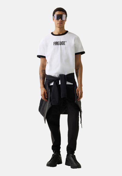 Ung mand iført store sorte futuristiske solbriller, hvid "FIRE&ICE" t-shirt, sorte bukser, bundet jakke og sorte sneakers, stående.