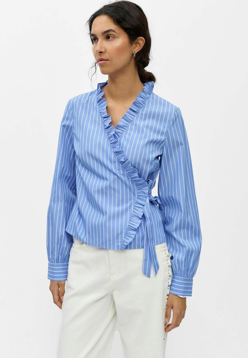 Femme portant un chemisier portefeuille rayé bleu et blanc avec un col à volants et un pantalon blanc avec un détail lacé sur le côté.