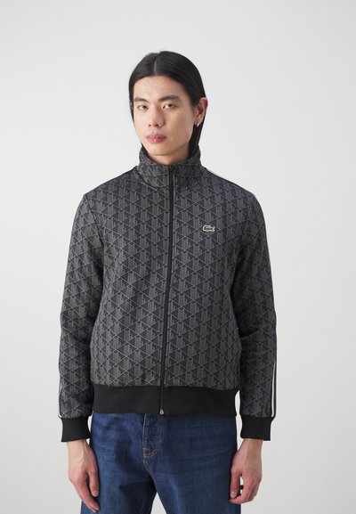 Veste zippée noire avec un motif géométrique, texture douce, col montant et logo de marque, associée à un jean bleu.
