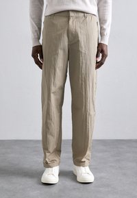 Beige, textured pants with a relaxed fit and elastic waistband, paired with white sneakers. Fabric appears lightweight and slightly crinkled.