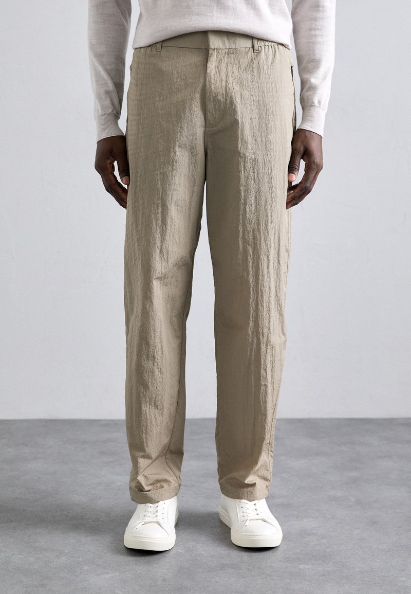 Beige, textured pants with a relaxed fit and elastic waistband, paired with white sneakers. Fabric appears lightweight and slightly crinkled.