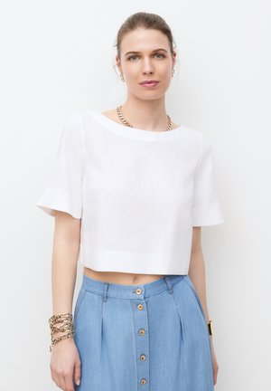 Jonge vrouw met haar naar achter gebonden, gekleed in een wit cropped topje, een blauw hoge taille rok met knopen aan de voorkant, gouden ketting, oorbellen en armbanden.