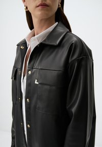 Giacca camicia in ecopelle nera con colletto a punta, dettagli in oro, tasche frontali e discreto logo sul petto.