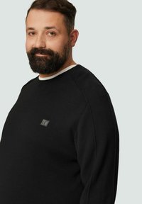 Schwarzer Pullover mit strukturiertem Strick und weißem Rundhalsausschnitt, mit einem kleinen schwarzen Logopatch auf der linken Vorderseite.
