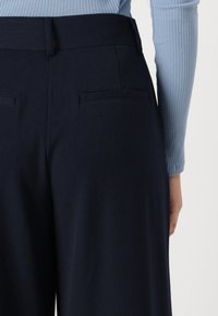 Navy high-waisted trousers with a smooth texture, featuring two back pockets and a wide-leg design. Complemented by a light blue ribbed top.