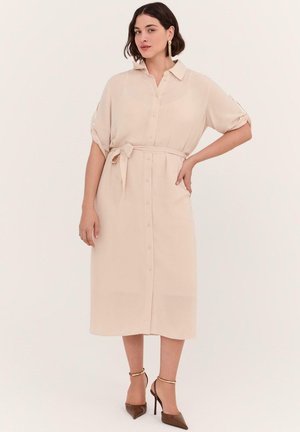 Kvinde iført beige midi-kjole med knapper, bindebånd i taljen og opkørte ærmer, kombineret med brune højhælede sko og guldringe.