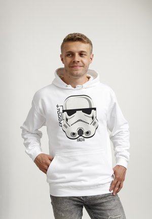 Ung mand iført hvid hættetrøje med sort og gråt Stormtrooper-hjelm-grafik, stående med hænderne i lommerne mod en ensfarvet baggrund.