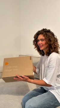 Caja de cartón en tonos neutros con el texto "Ámame. Llévame puesto." en el frente. Sostenida por una persona sentada sobre una superficie texturizada de color claro.