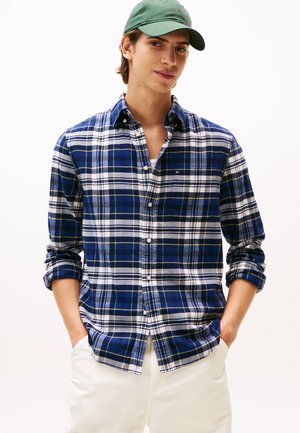 Camisa de franela a cuadros azul y blanca con acentos amarillos, cuello abotonado y mangas enrolladas; presenta un pequeño logo en el pecho.