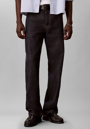 Mann trägt dunkle Straight-Leg-Jeans, schwarzen Gürtel, schwarze Ledermokassins, ein silbernes Armband und ein blau-weiß gestreiftes Hemd.