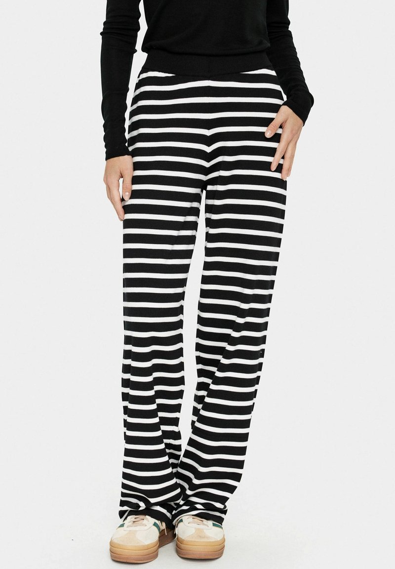 Personne portant un pantalon large à rayures horizontales noir et blanc, un haut à manches longues noir et des baskets blanches avec des semelles en gomme.