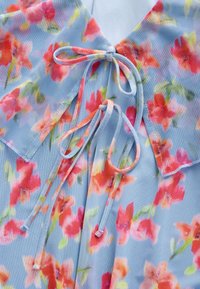 Florale Bluse aus hellblauem Stoff mit lebendigen pinken und orangen Blumenmustern, einem Kragen und einer Schleifenverzierung vorne.