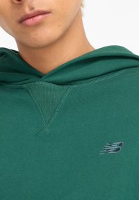 Felpa verde con cappuccio a trama e piccolo logo grigio "New Balance" sul petto. Tessuto morbido e liscio con dettagli a costine vicino al collo.