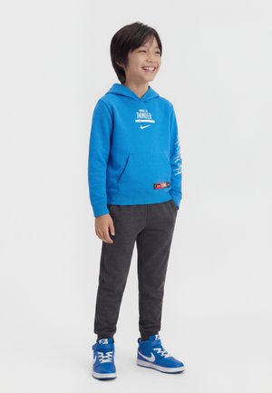 Garçon souriant, portant un sweat à capuche bleu Oklahoma City Thunder, un pantalon de jogging foncé, et des baskets Nike bleues, debout avec les mains dans les poches.