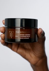 Pot en verre ambre avec un couvercle noir, étiqueté "True Botanicals Calm Ginger Turmeric Cleansing Balm" en texte blanc. Surface lisse, bords arrondis.
