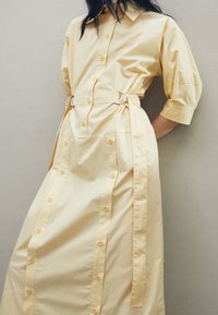 Ljusgul skjortklänning med korta puffärmar, knäppning fram, avtagbart bälte med silverdetaljer och sidofickor. Slät tygkvalitet.