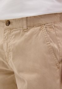 Pantaloni corti in cotone beige con chiusura frontale a bottone, passanti per cintura e un piccolo dettaglio di logo ricamato di lato. Tessuto liscio.