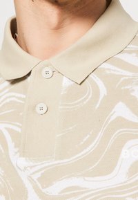 Gros plan d'un polo marbré beige et blanc avec deux boutons et un col plié sur le cou et la poitrine supérieure d'une personne.