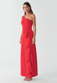 Vestido maxi rojo de un solo hombro con un cuerpo ajustado y detalles de volantes a lo largo del lateral. Hecho de tela suave y ligera.