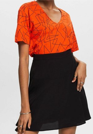 Femme portant un t-shirt orange à col en V avec des lignes abstraites noires et une jupe noire taille haute, avec des bagues et un bracelet en argent.