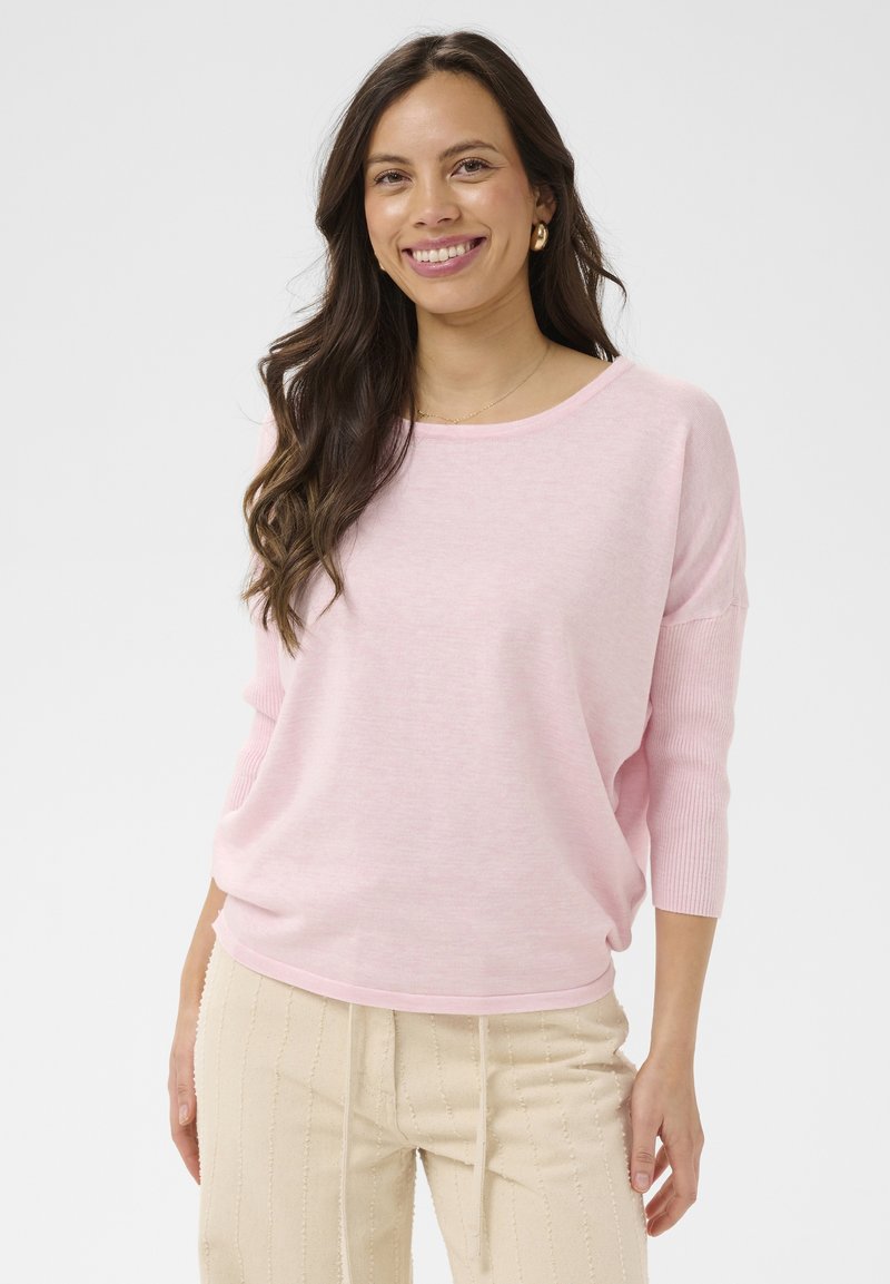 Femme souriante aux longs cheveux bruns, portant un pull rose clair et un pantalon beige rayé, debout devant un fond blanc uni.