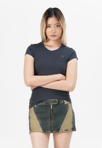 T-shirt blu navy a maniche corte con scollo tondo e logo; abbinato a una minigonna di jeans con zip caratterizzata da pannelli verdi e beige.
