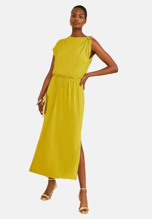 Frau trägt ein ärmelloses senfgelbes Midikleid mit Schulterdetail, hält eine beige Clutch und trägt goldene Riemchensandalen.